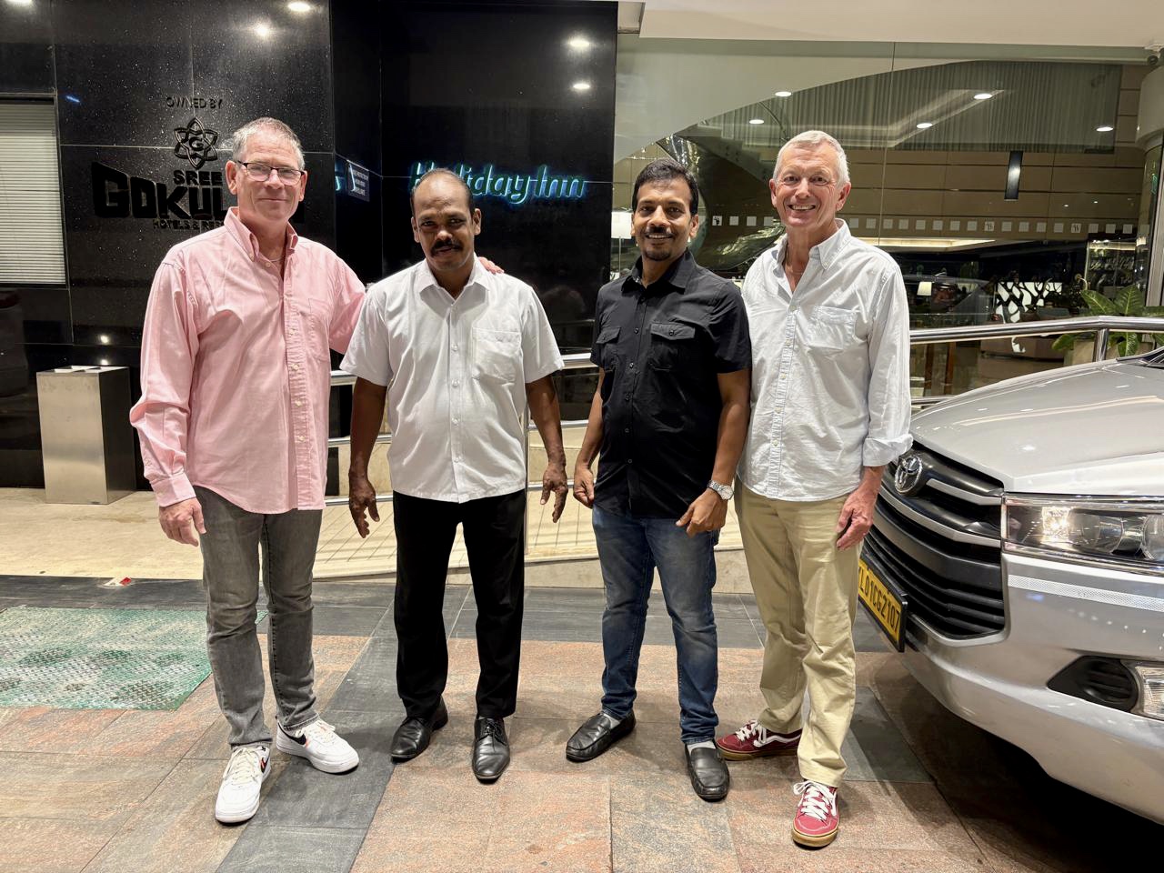 Four men pose together indoors beside a parked white SUV, smiling at the camera near a building entrance.
