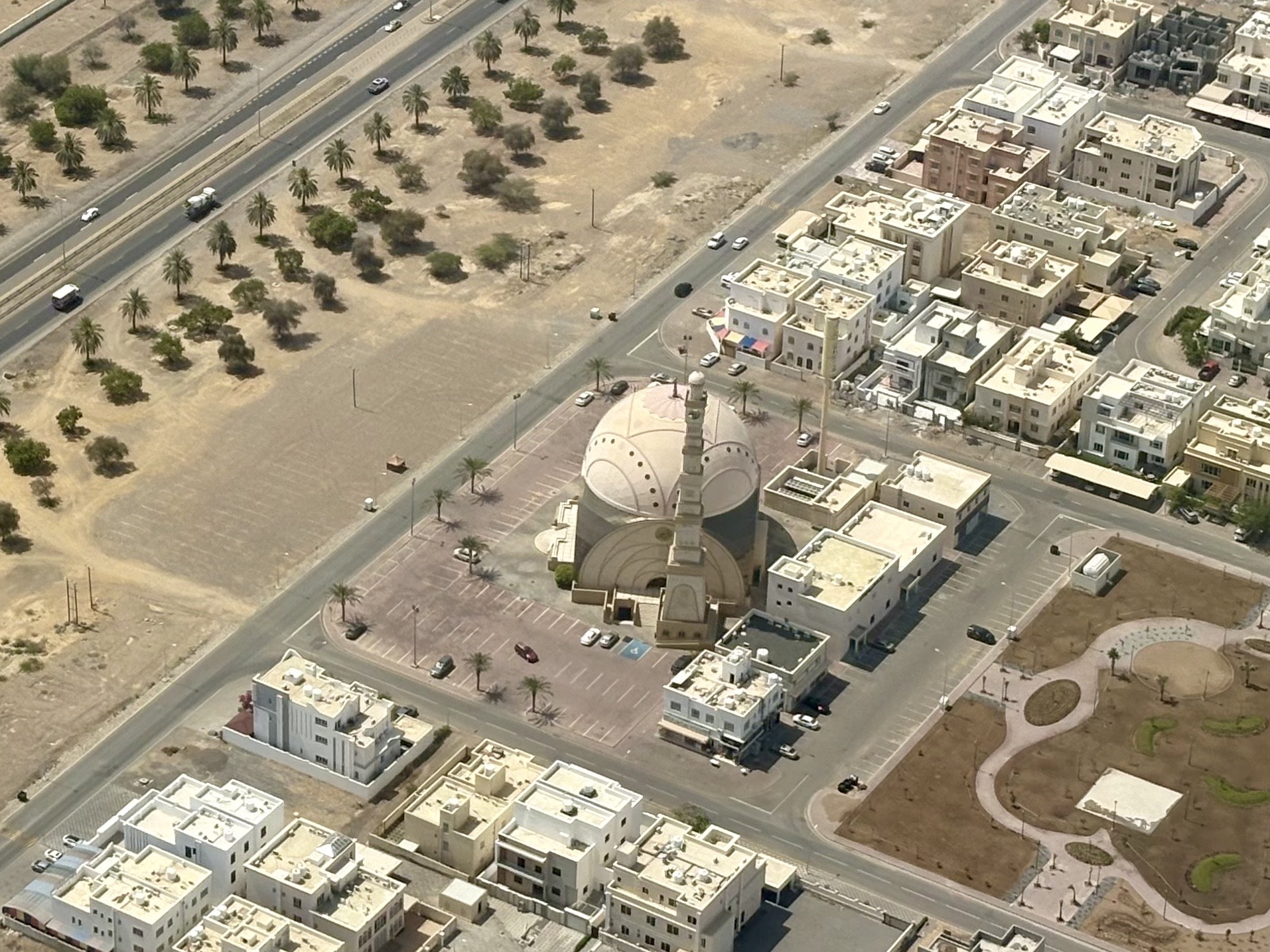 Aerial view of a desert city with a large domed mosque and minaret next to beige apartment buildings and palm-lined streets around parking lots.
