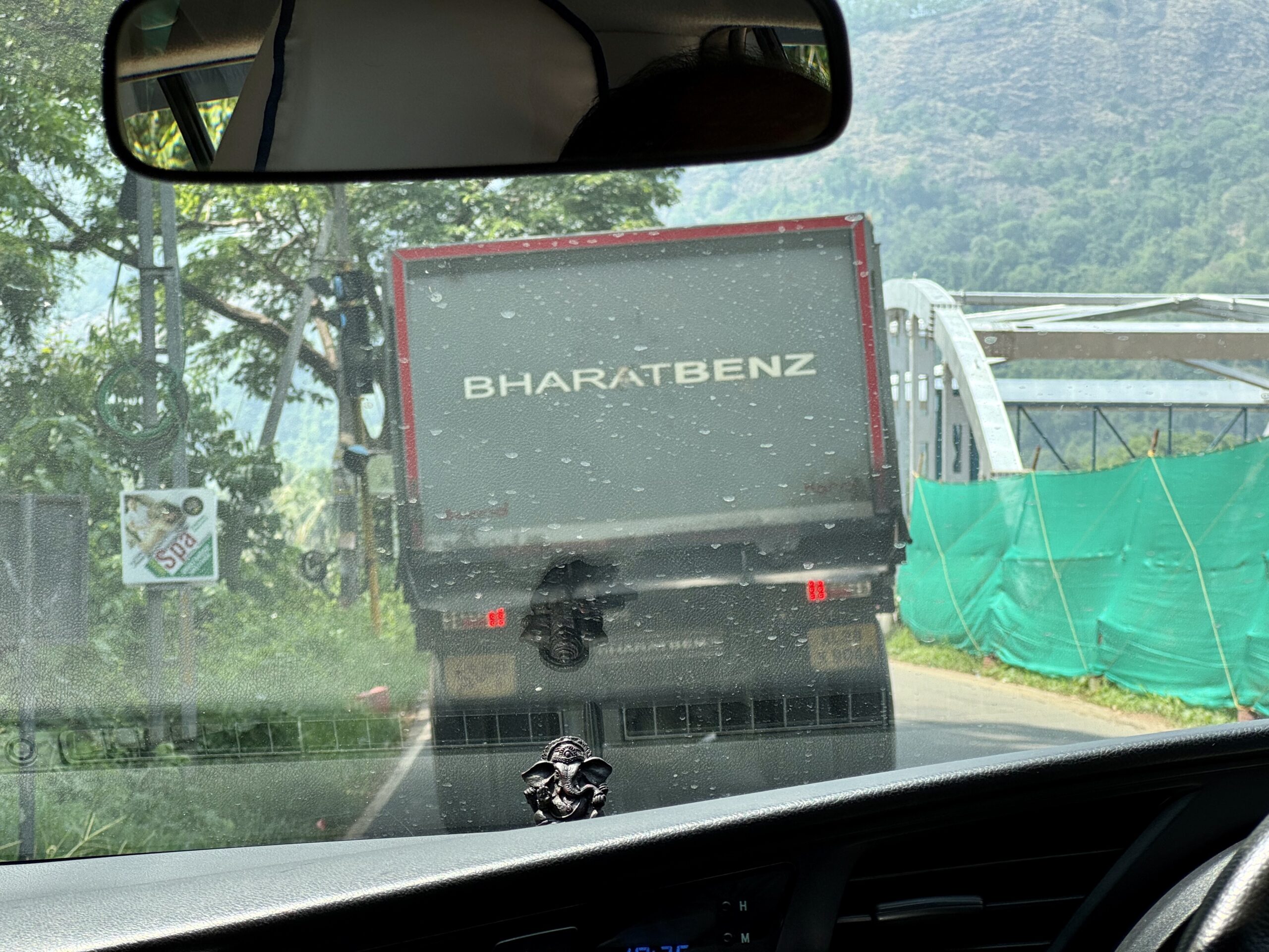 View from inside a car: a BharatBenz truck in front, raindrops on the windshield, construction fencing on the right, and a rearview mirror overhead.