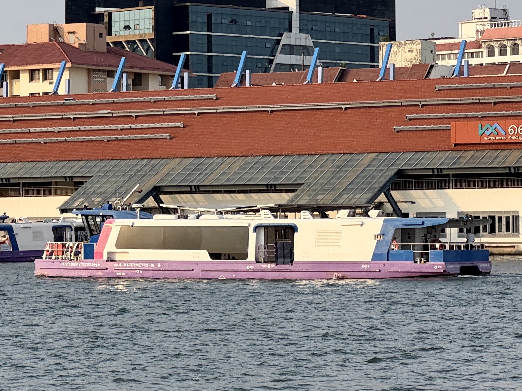 Purple and white passenger boats docked at a marina in front of a red brick terminal with blue diagonal supports and a Water Metro sign visible on the building.