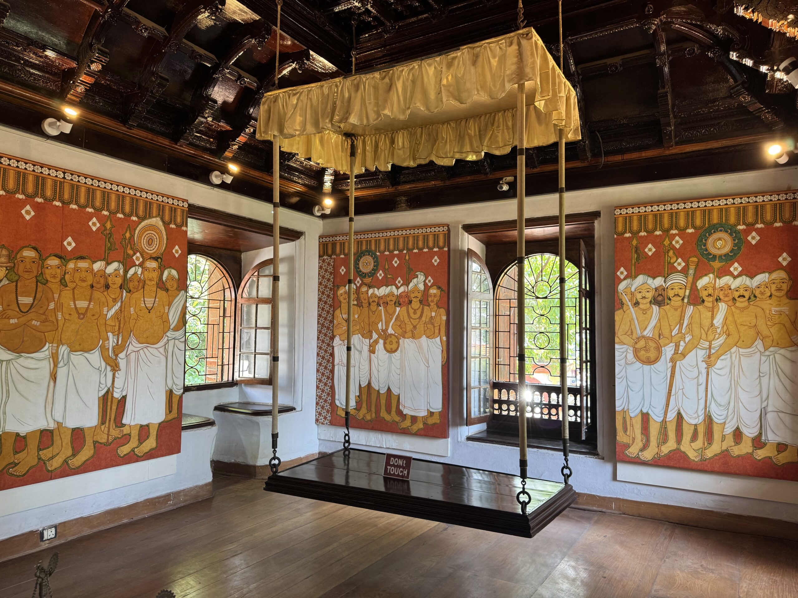 Large wooden swing suspended from the ornate ceiling, enclosed by colorful mural panels of monks in orange robes around a gallery room.