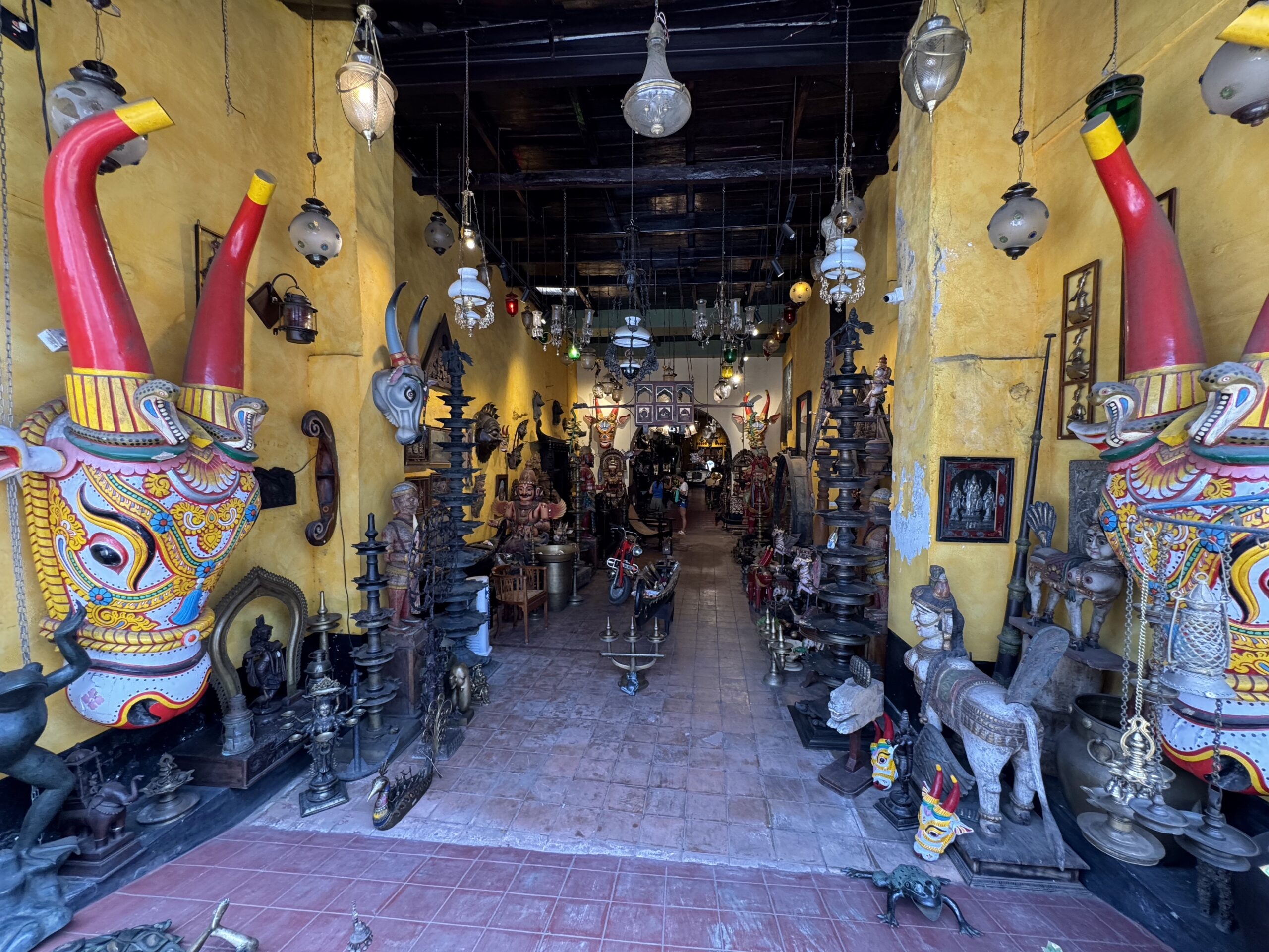 Colorful shop interior filled with large painted statues and ornate carvings along yellow walls, with hanging lamps overhead.