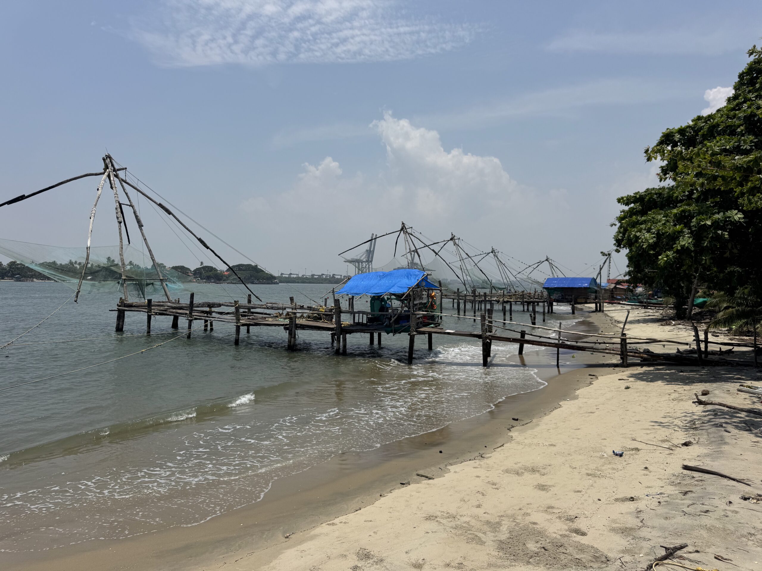 Beached fishing platform with wooden poles and nets in shallow water, blue tarps over small huts along the shore, and a tree line on the right.