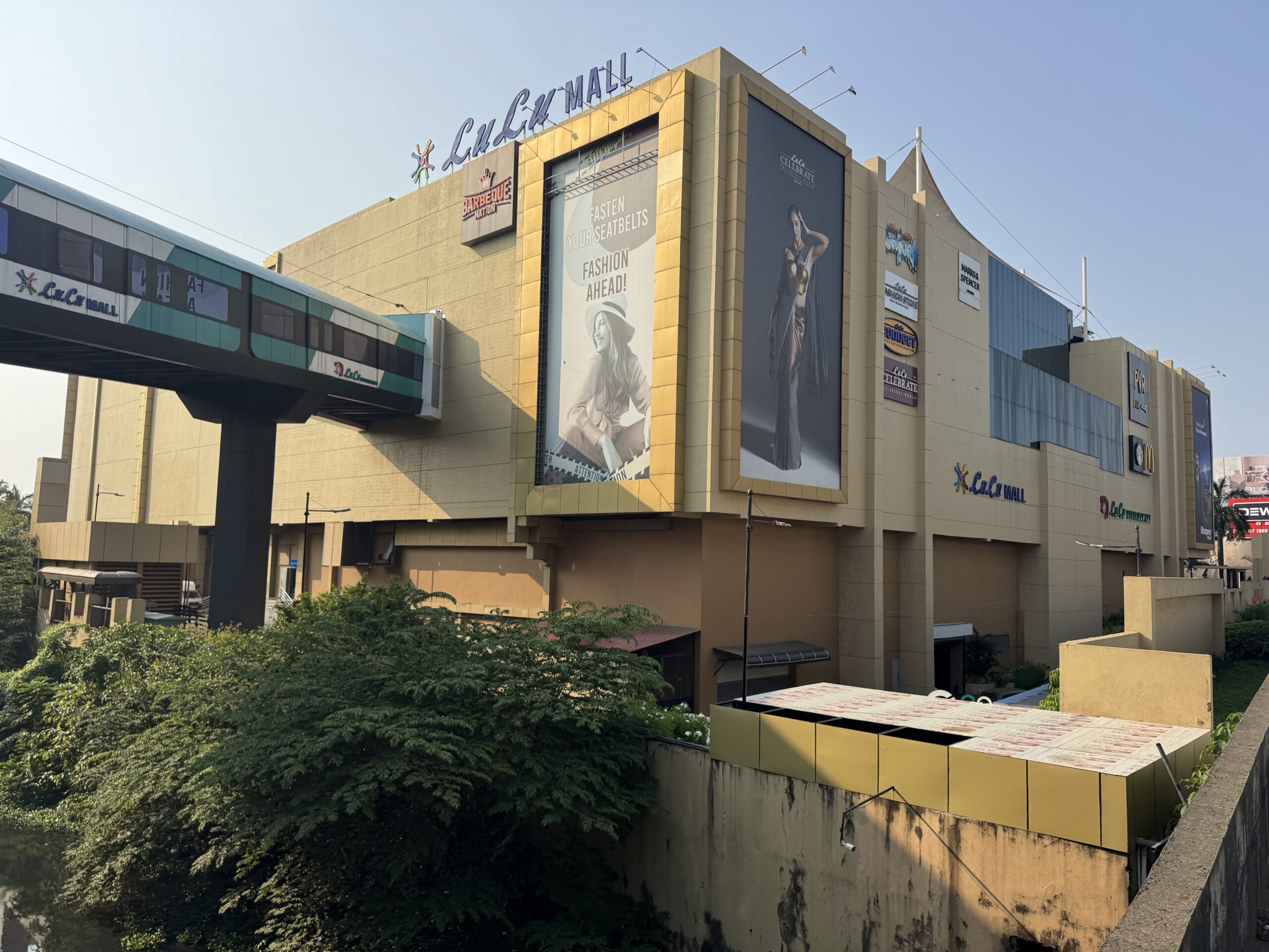 Exterior view of a shopping mall with large advertisement banners on the façade and a suspended monorail passing by on a blue sky day, with greenery in the foreground, creating urban contrast.