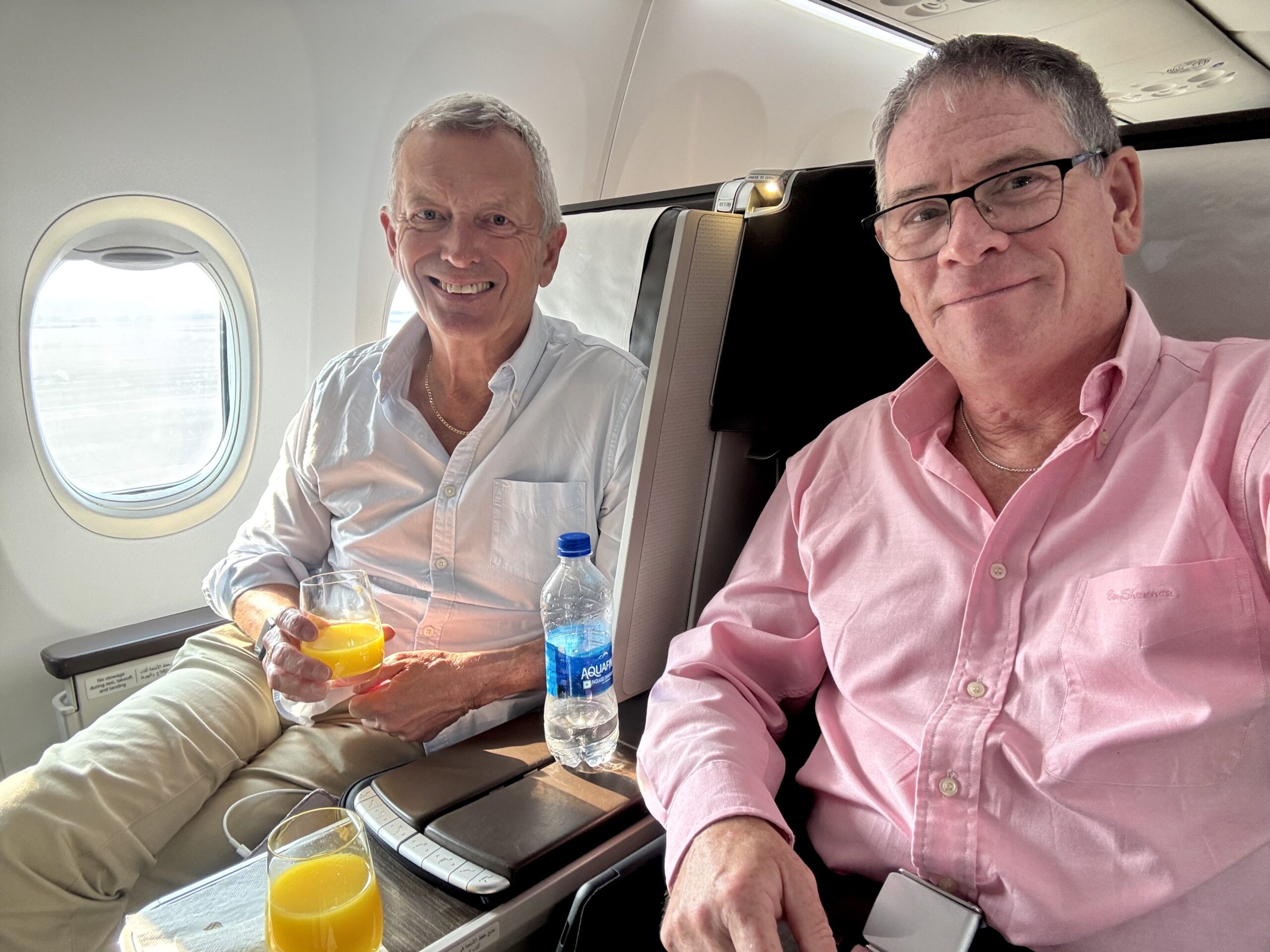 Two men sit in business-class seats on a plane, smiling, with orange juice and a water bottle on the tray in front of them.