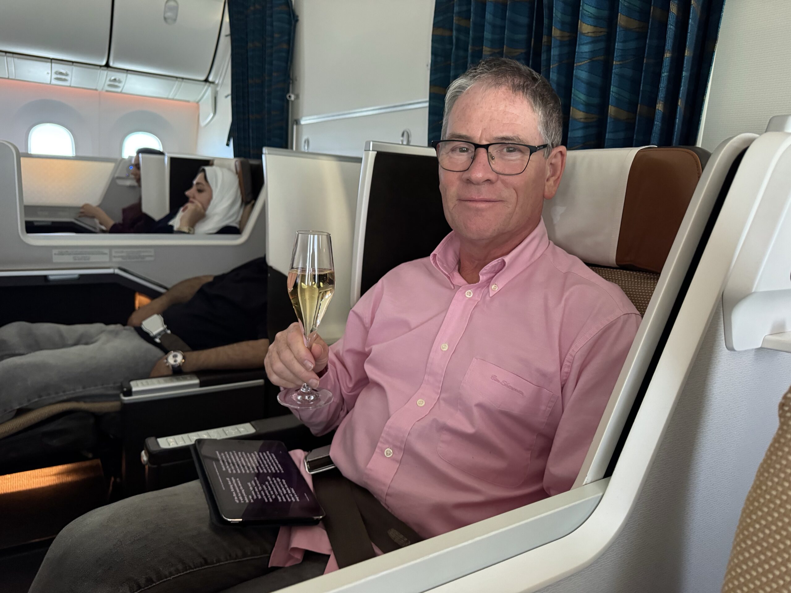 Man in a pink shirt and glasses sits in a business-class seat, holding a champagne flute toward the camera with a smile.