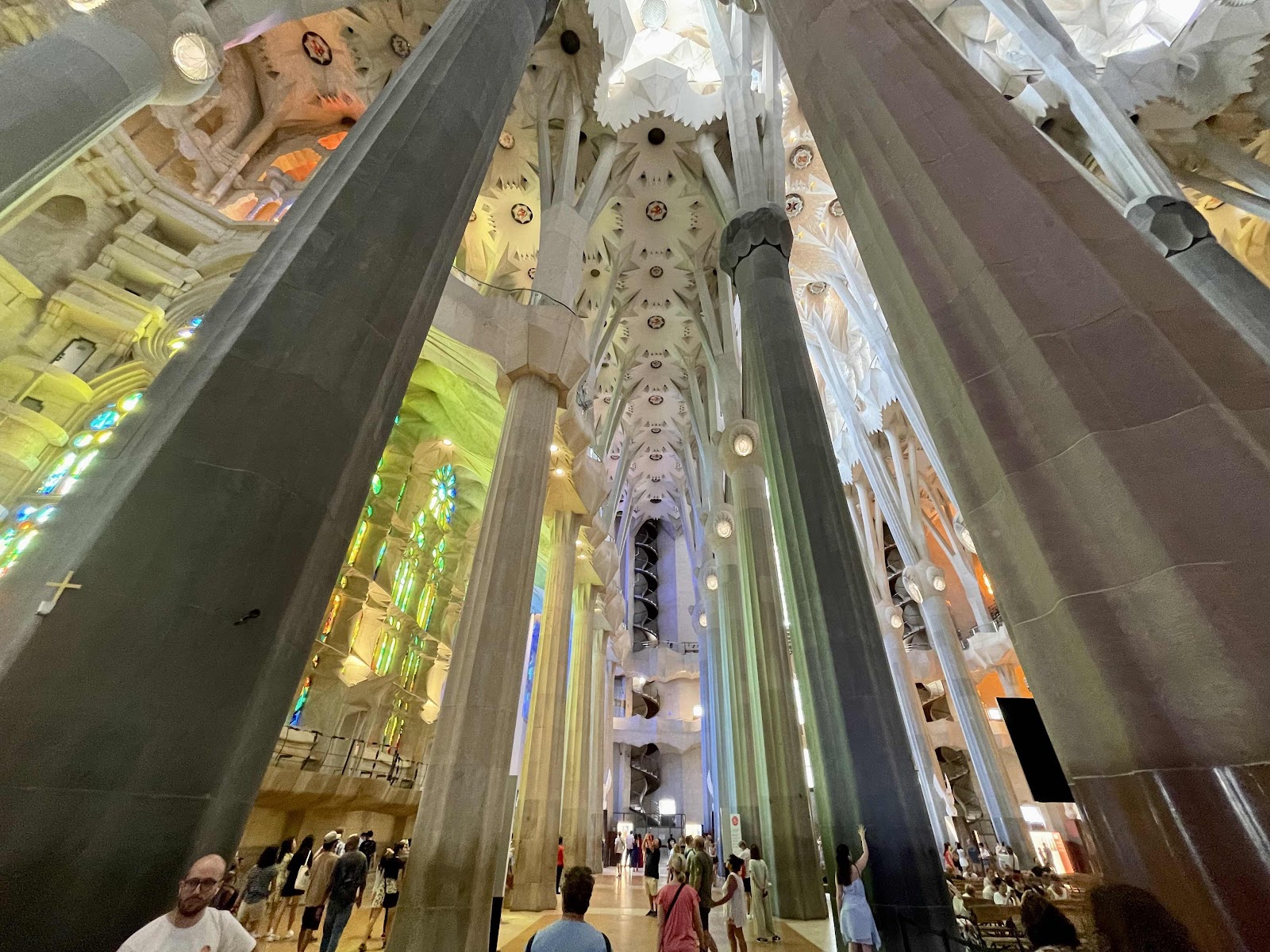 Inside the Sagrada Família's nave: towering tree-like stone columns rise to a colorful, geometric ceiling with stained-glass light. Visitors walk the vast interior.