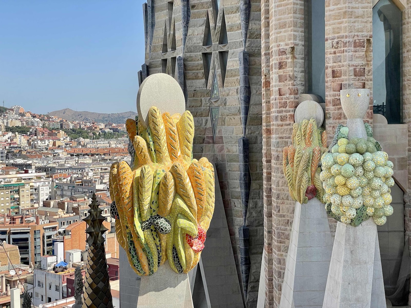 Colorful mosaic cactus sculptures on the exterior of the Sagrada Família, with Barcelona cityscape in the distance.”
