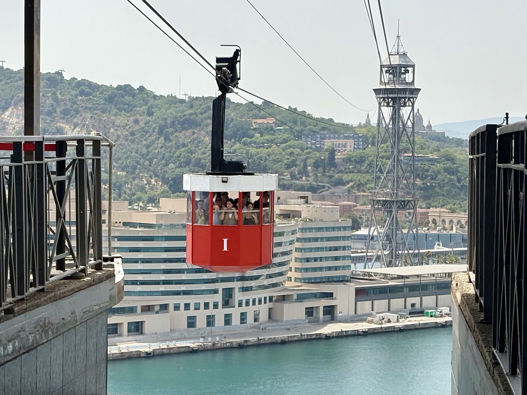 Red gondola carriage with passengers suspended over a waterway, moving toward a city skyline and a tall tower in the distance.