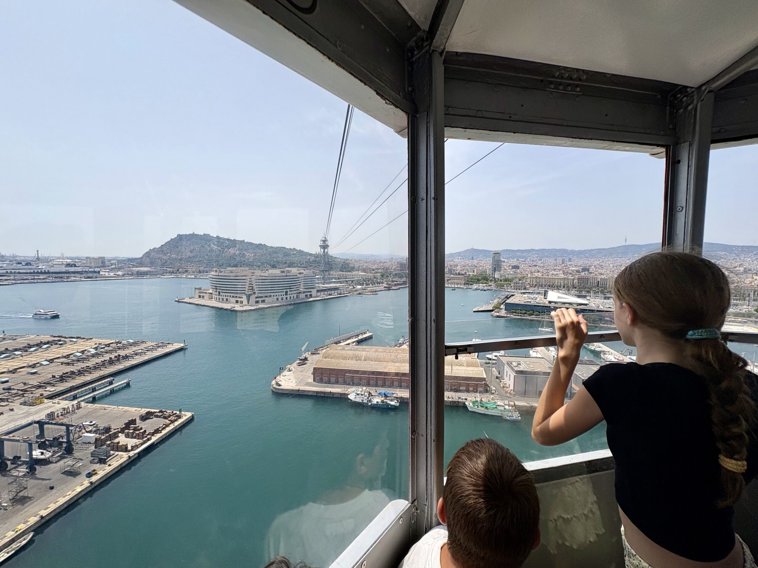 Passengers look out from a glass-walled cable car at a harbor, with docks, water, and city skyline in the distance.