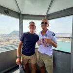 Two men posing inside a glass-observation capsule with a city harbor below on a sunny day.