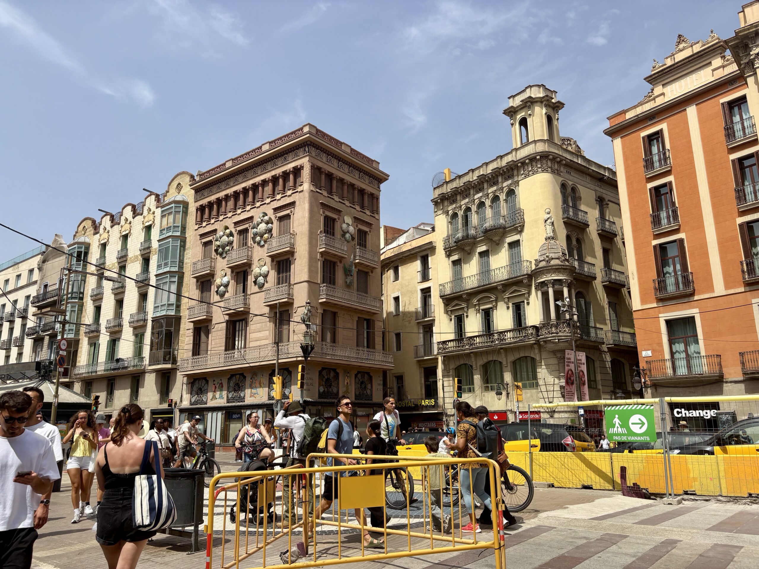 Busy city street with ornate European buildings, pedestrians and cyclists behind yellow barricades on a sunny day in front of storefronts and balconies.