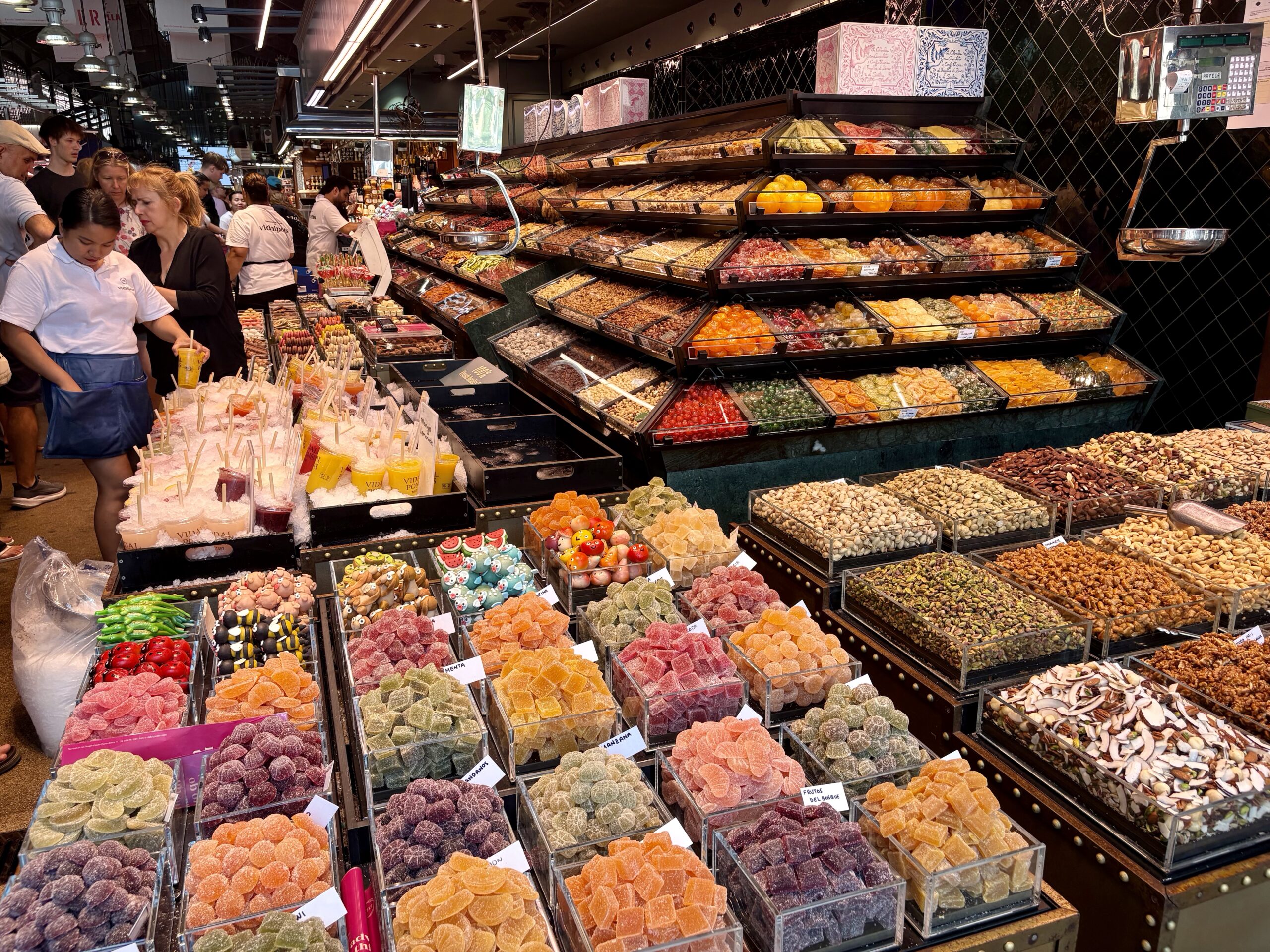 Colorful display of candies in a busy market shop with customers browsing in the background.