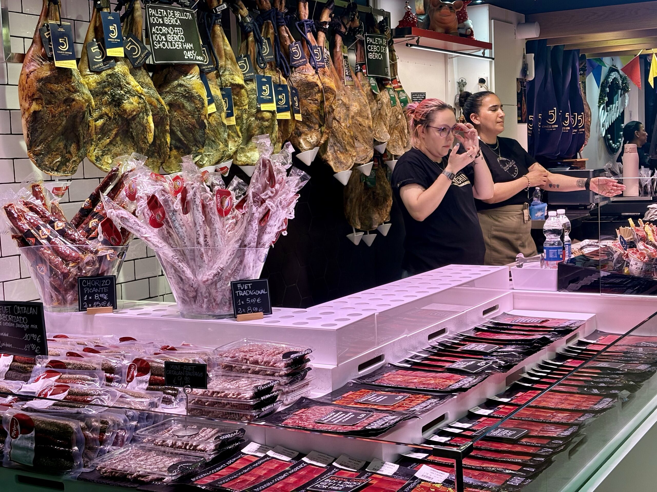 Deli counter with hanging cured hams and price tags, sausages wrapped in plastic, and two staff members behind the counter