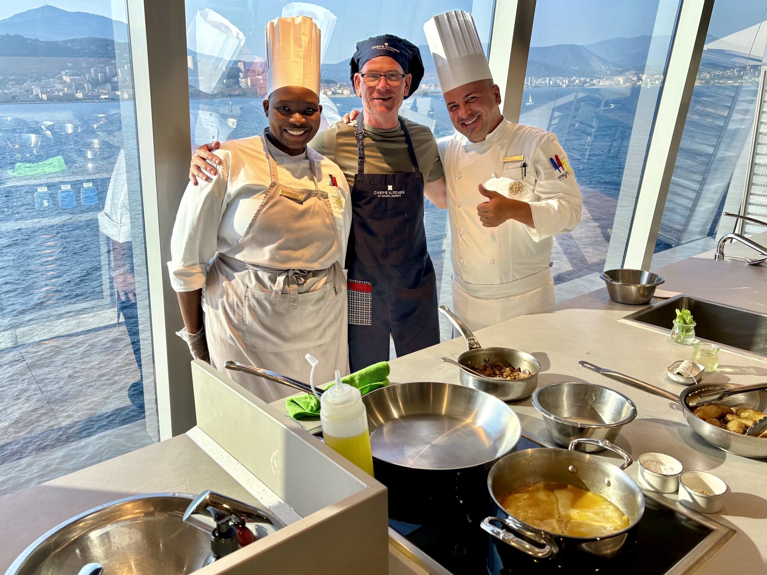 Three chefs pose together in a bright shipboard kitchen, wearing tall hats and aprons, with pots and bowls of food in front.