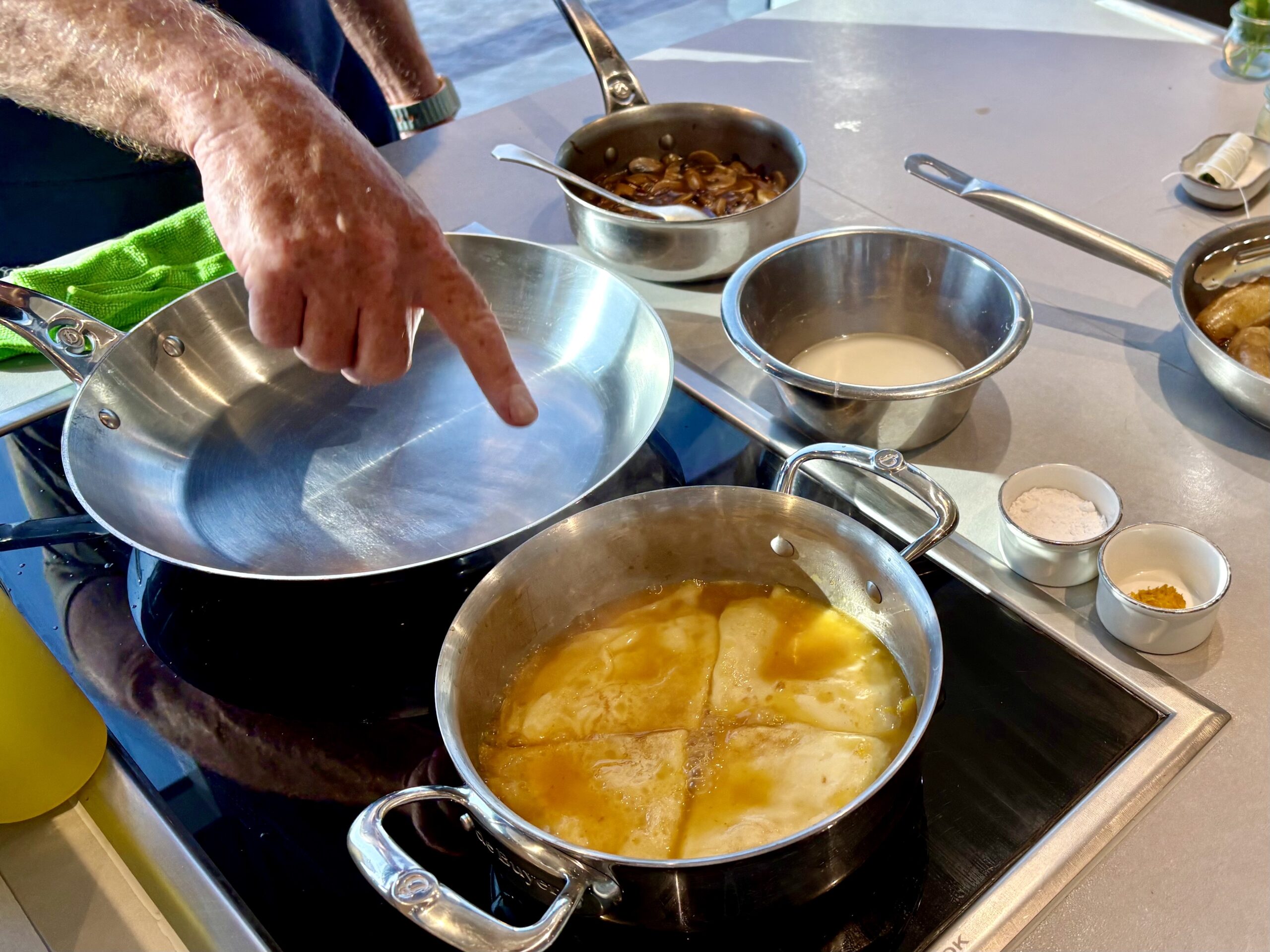 A cook’s hand points toward a large stainless steel pan with bubbling sauce on a kitchen stove, surrounded by other pots and utensils.