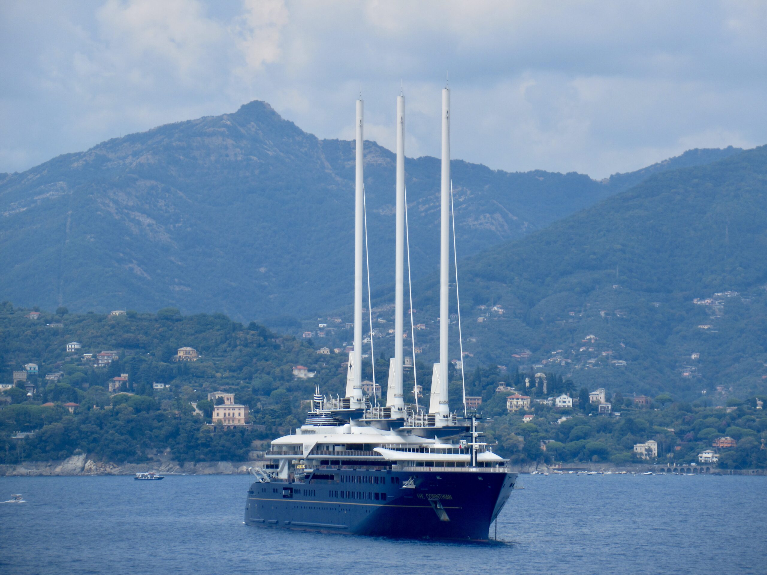 Large cruise ship with four tall masts docked in a bay, with hills and a town in the background.