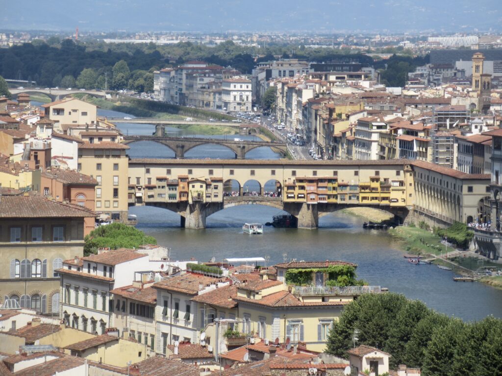 Aerial view of a European city with red-tiled rooftops, a river, and stone bridges across the water, under a blue sky.