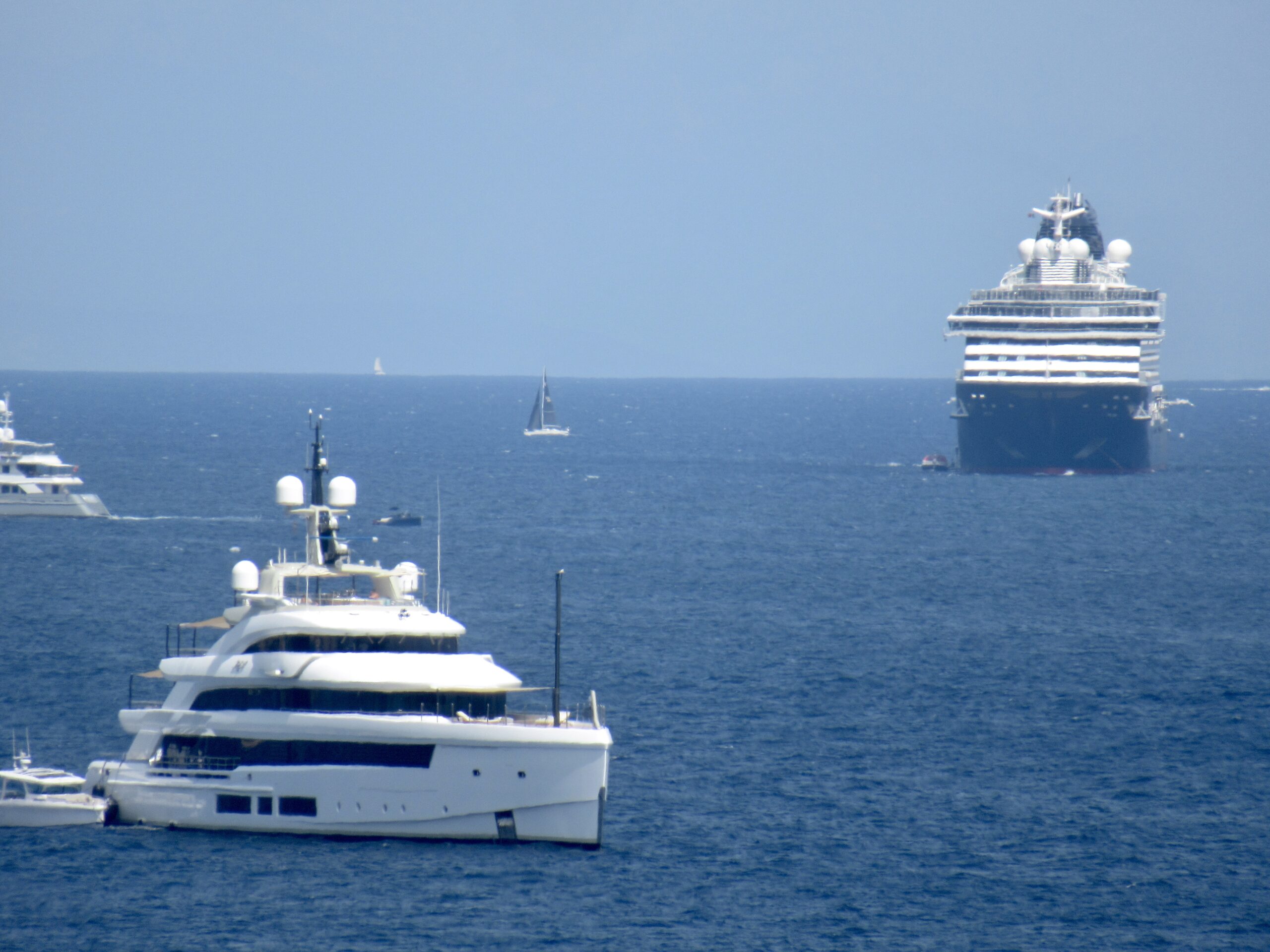 Large white cruise ship on the open sea with several smaller boats nearby under a clear blue sky.
