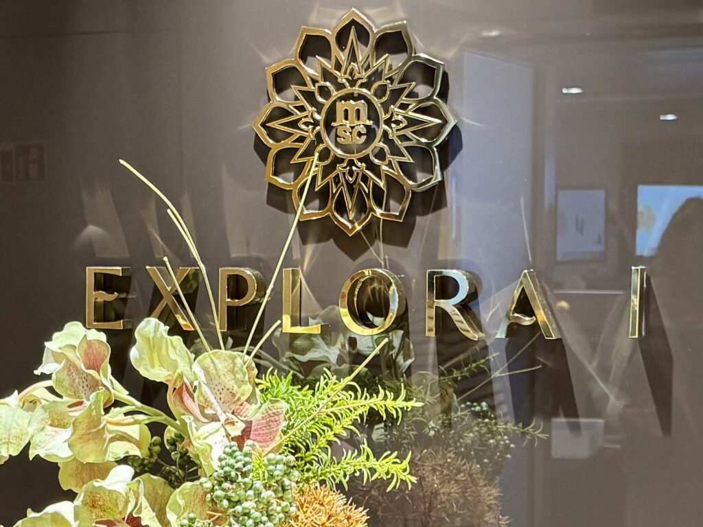 Gold letters spell EXPLORAI in a storefront window, with a gold ornamental emblem above and a floral arrangement below.