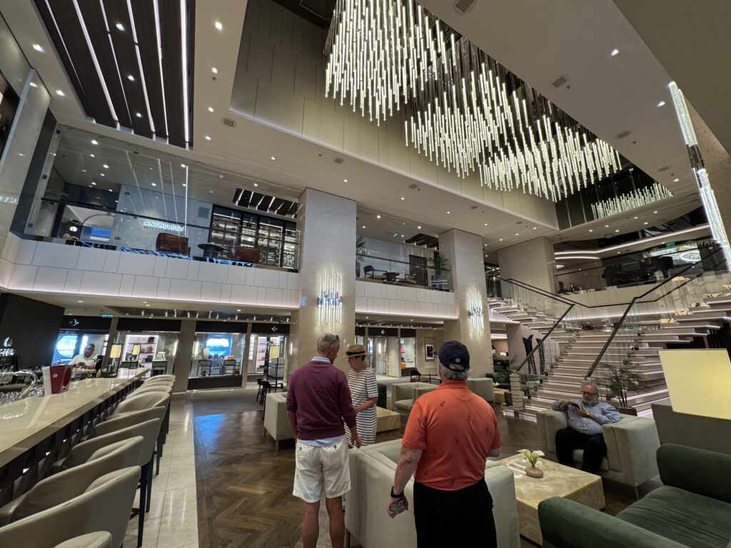 Luxurious hotel lobby with tall glass windows, curved staircase, and a dramatic ceiling installation of hanging vertical light rods; several people stand and chat.