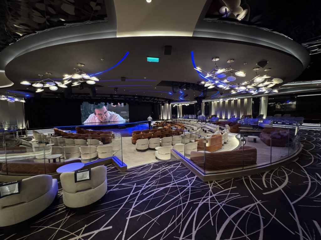 Modern auditorium with curved beige seats, glass railings, blue neon lines, and a large stage displaying a man on screen.