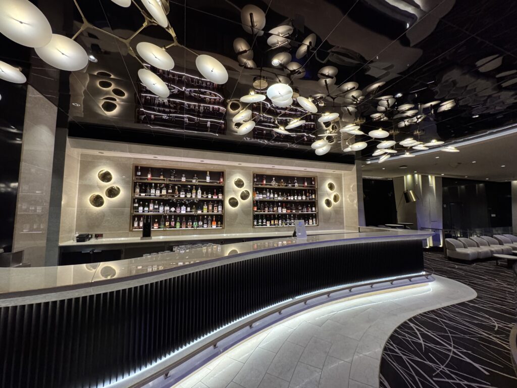Modern bar interior with a long black counter, backlit liquor shelves, and a suspended installation of white circular discs on the ceiling.