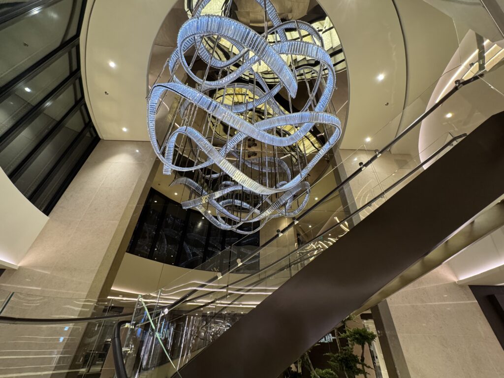 Twisting illuminated sculpture with looping blue ribbons hanging in a multi-level atrium surrounded by glass railings and beige walls.