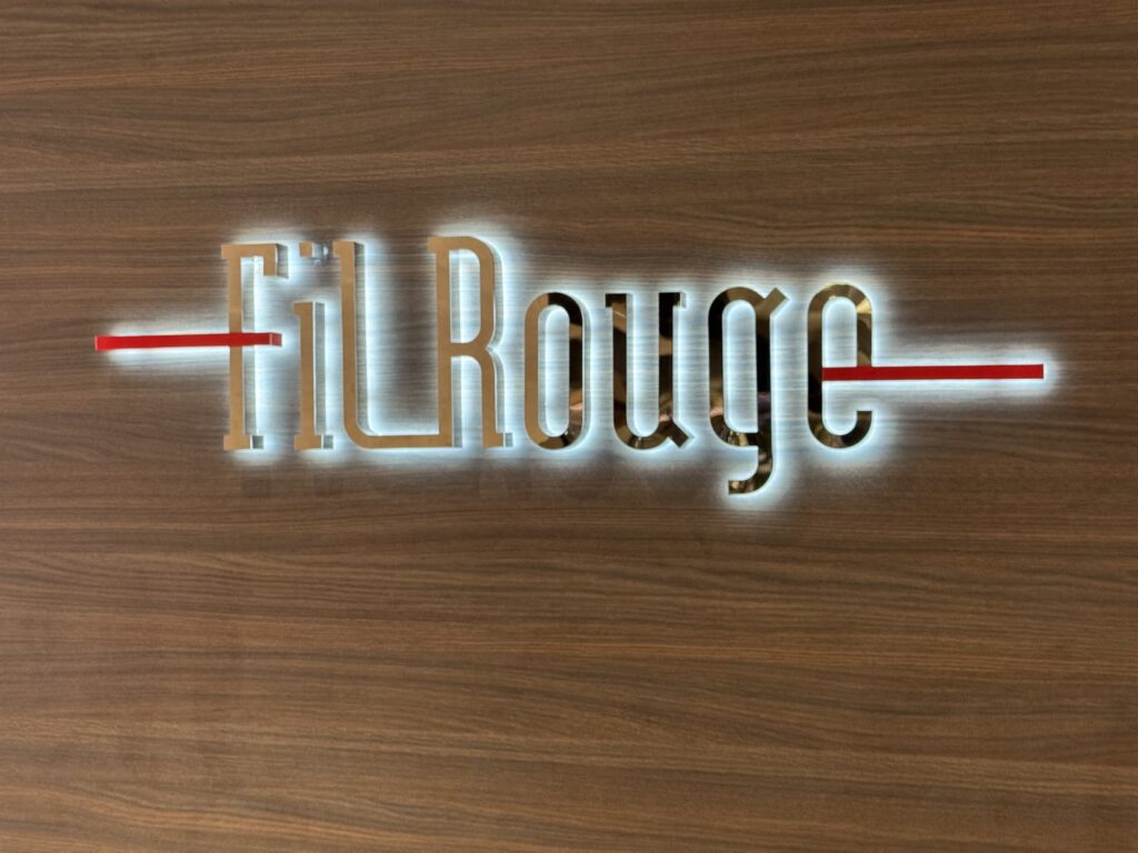 Backlit metal sign with stylized letters mounted on a wood wall, featuring two red horizontal bars crossing the sign.