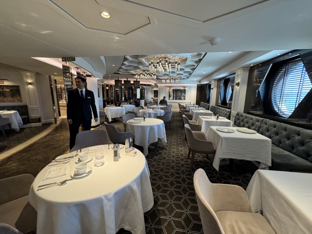Elegant dining room with round tables, white tablecloths, and beige chairs, chandeliers overhead and a waiter in a suit nearby.