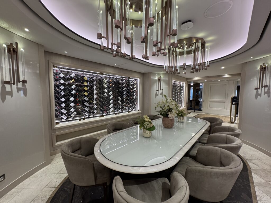 Luxurious wine lounge with a wall of bottles, glass dining table, and geometric chandeliers above.