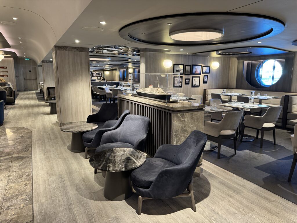 Upscale dining room with gray chairs, round marble-topped tables, and a ship model display on a marble counter center stage.
