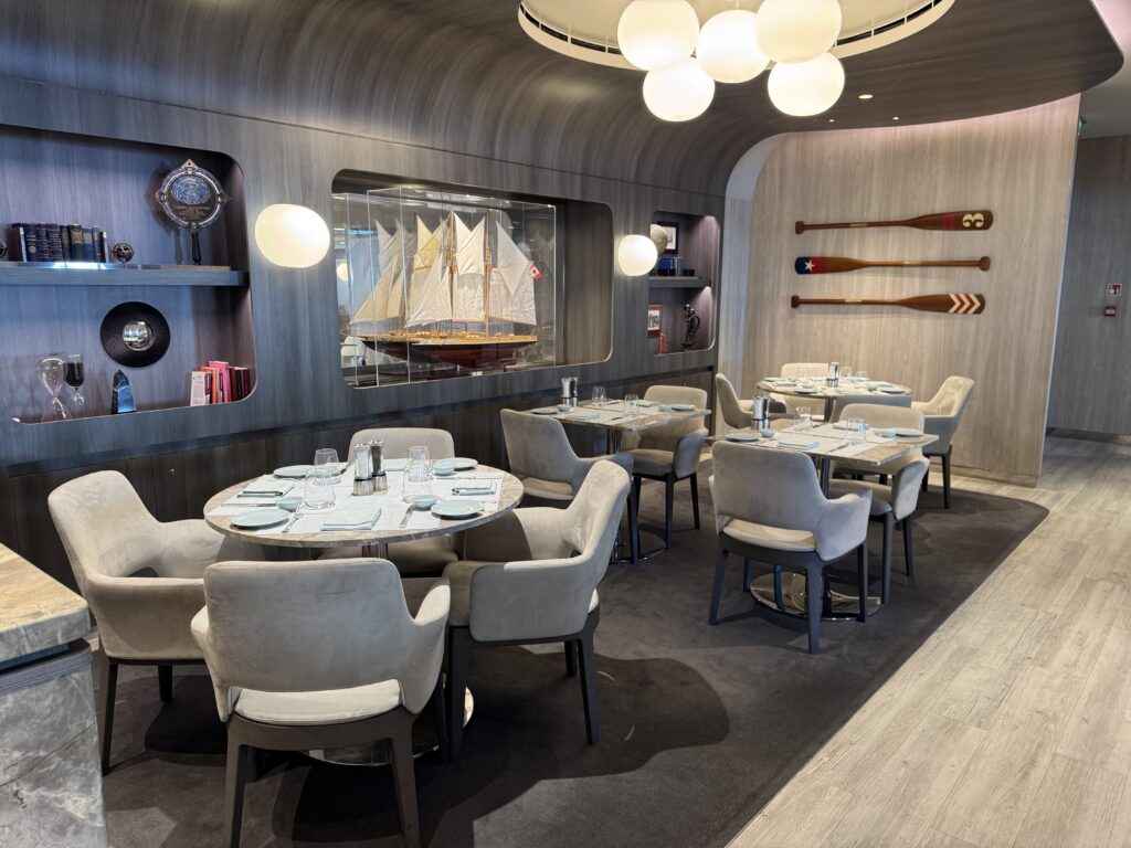 Restaurant dining area with round tables set for guests, beige upholstered chairs, wall-mounted oars, and a ship model display behind glass in a nautical-themed room.