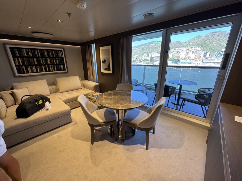 Cruise ship lounge with a beige sectional sofa, round marble table, and large windows looking out at water and distant mountains.