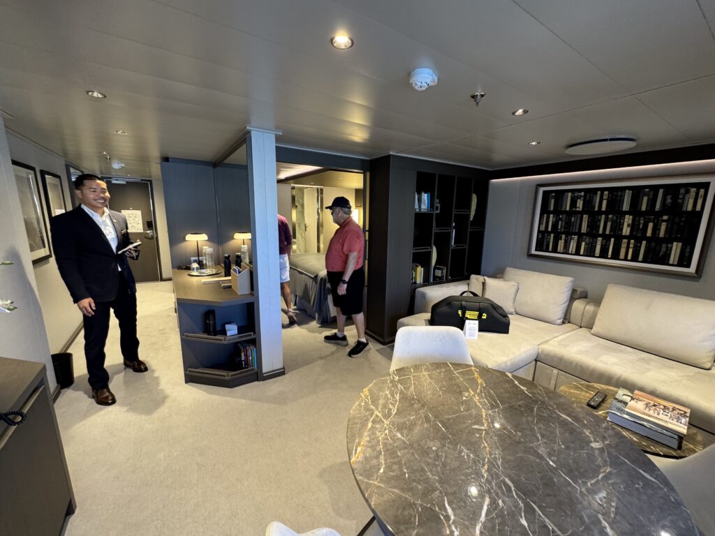 Interior living area on a yacht or modern lounge: gray sofa, marble table, built-in shelving, and two men standing with one in a suit and the other in a red shirt.
