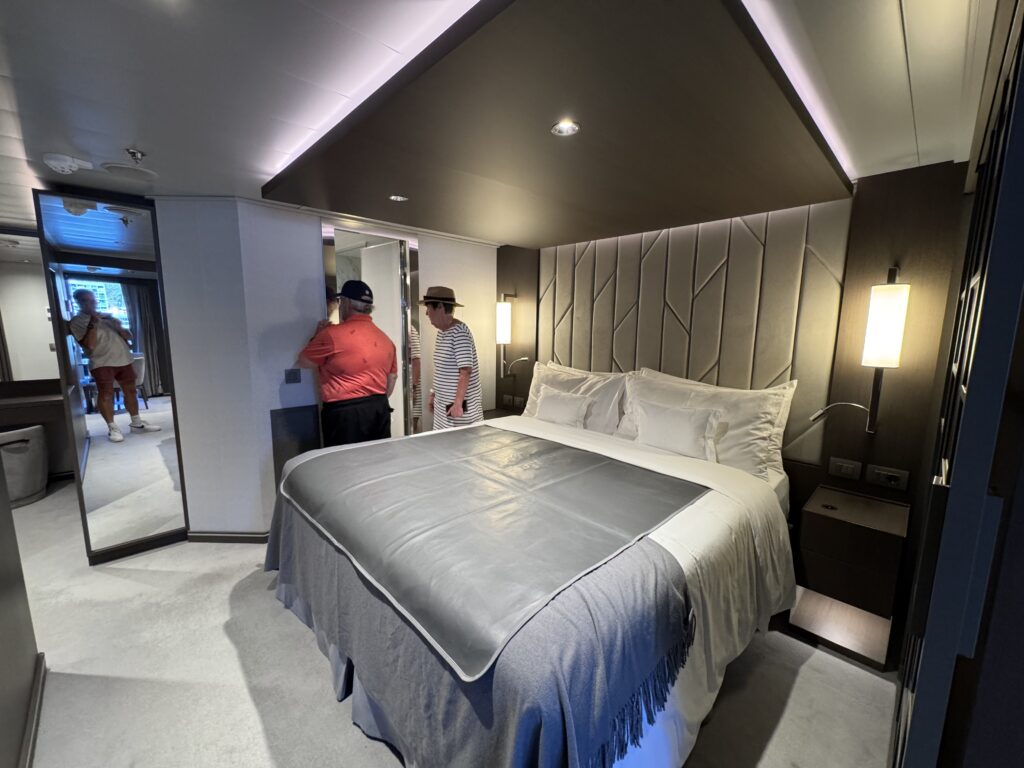 Contemporary hotel room with a large bed, gray bedding and a padded headboard; two guests stand near a mirror in the background.