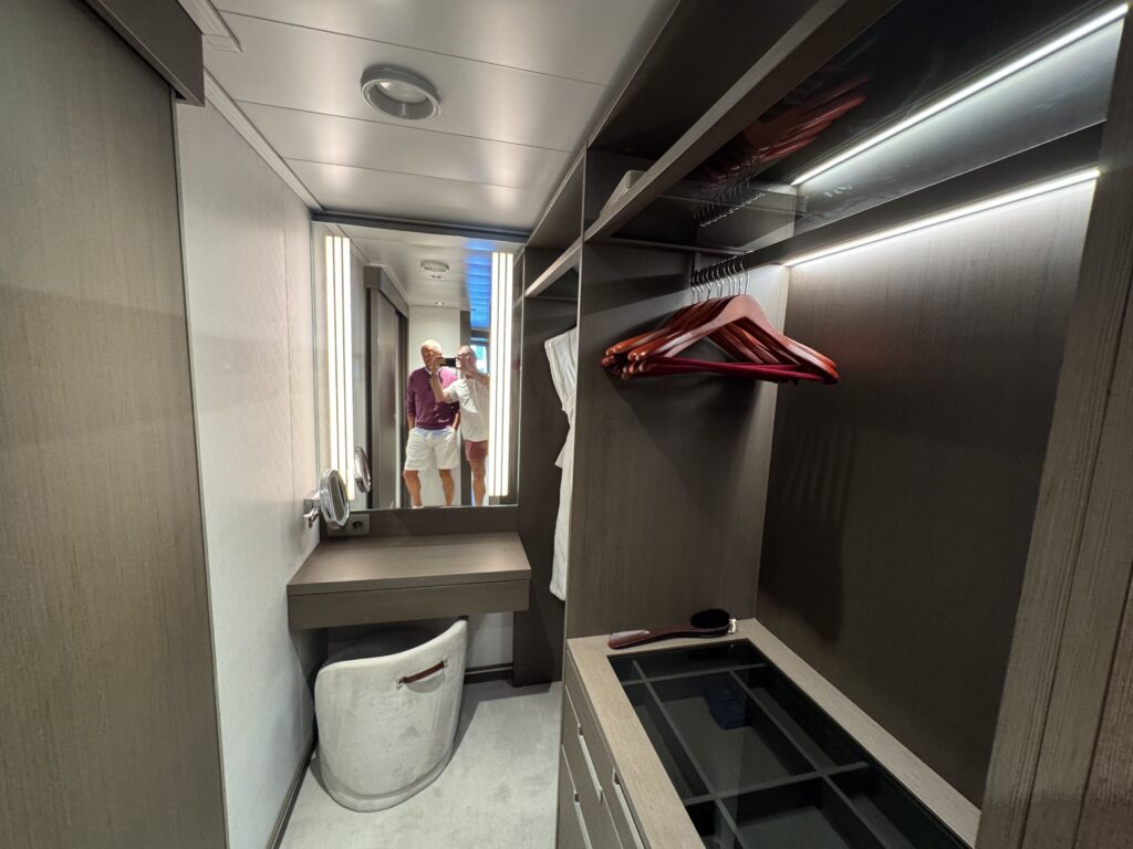 Walk-in closet with dark wood shelving, hanging red clothes, a full-length mirror, and two men taking a photo in the reflection.