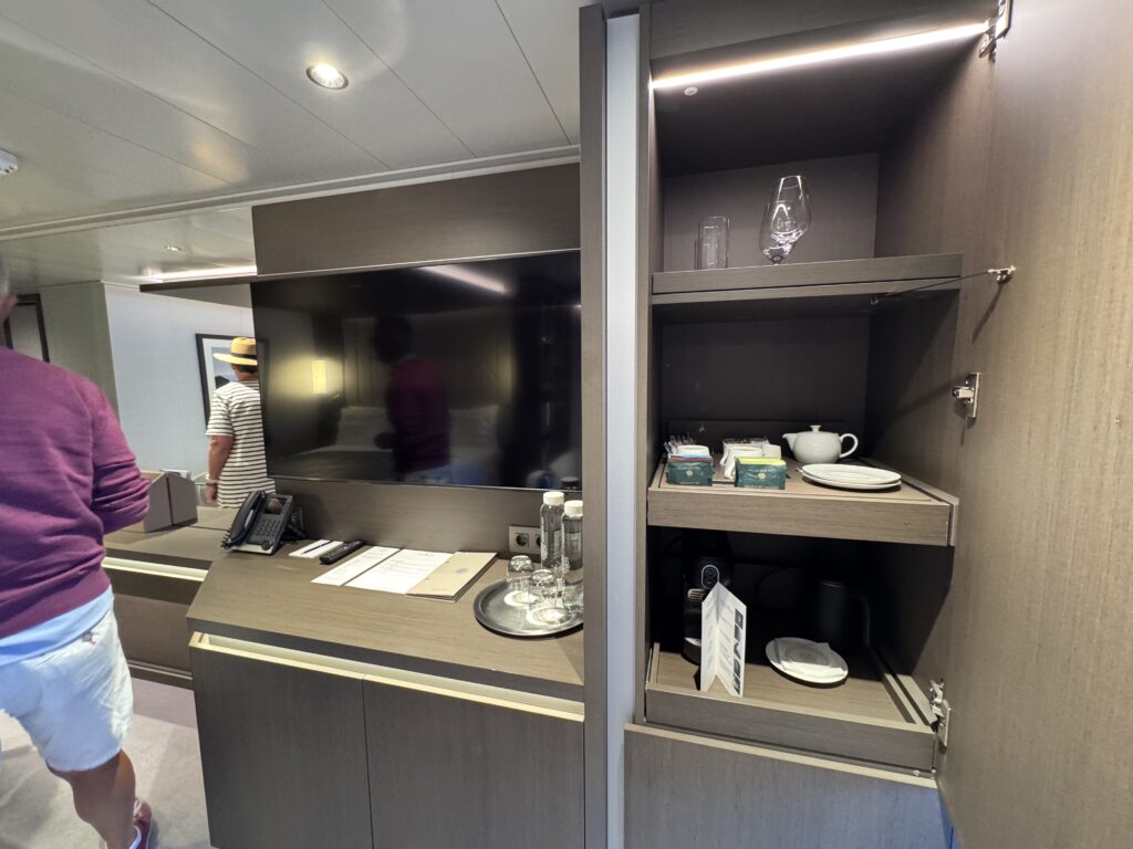 Showroom kitchen cabinet with glassware on the top shelf, a teapot and cups on the lower shelf, and a person in a purple shirt at the counter.