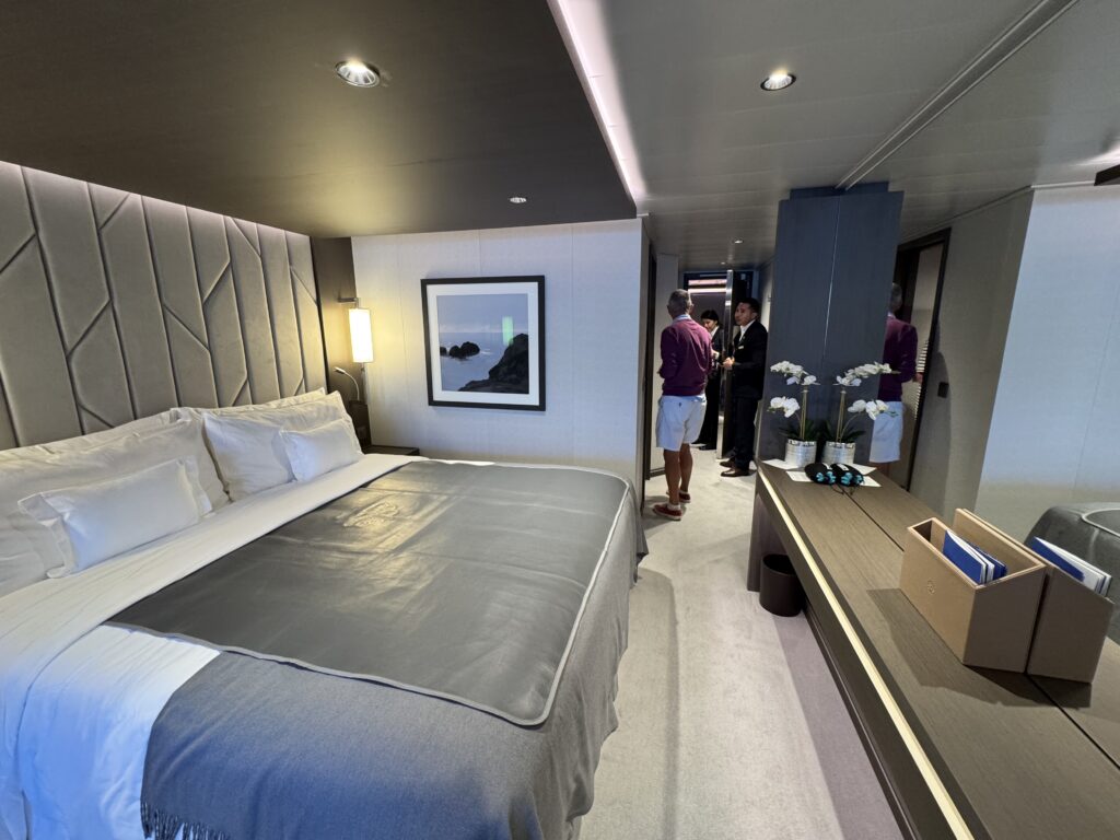 Hotel bedroom with two large beds and gray-blue bedding, modern padded headboards, framed sea landscape on the wall, and a desk with orchids; people stand near an open elevator in the background.