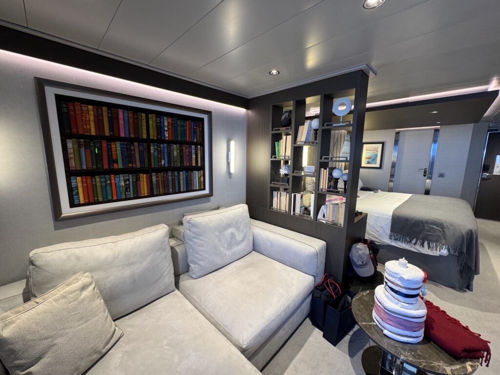 Interior of a modern yacht lounge with a light gray sectional sofa and a dark wood shelving unit nearby.