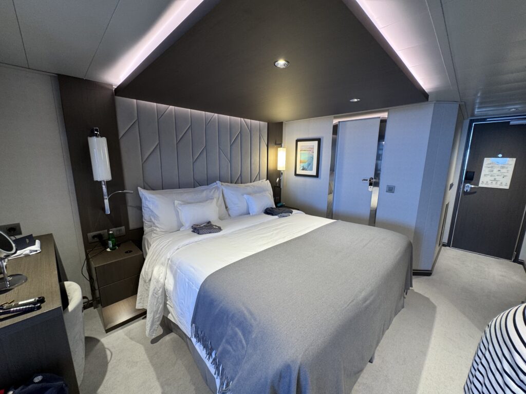 Modern hotel room with a large bed, white linens, and a tufted gray headboard wall with angled panels.
