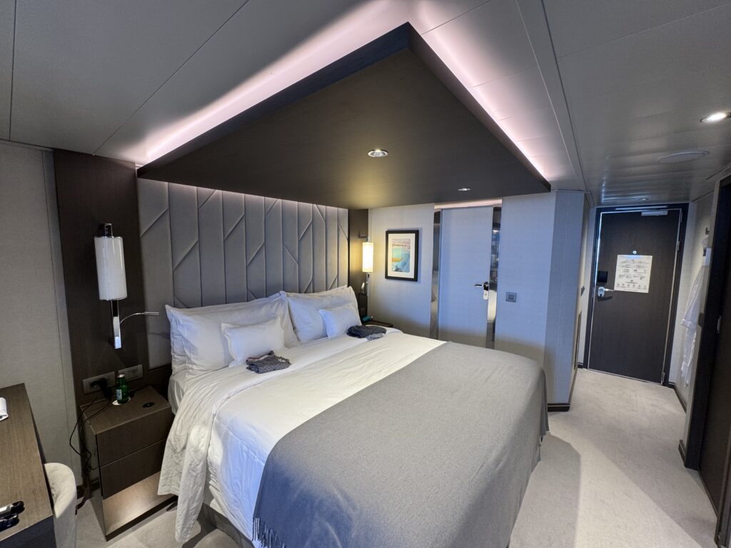 Hotel bedroom with a large bed, padded headboard, and soft lighting; doorway and framed art visible in the background.