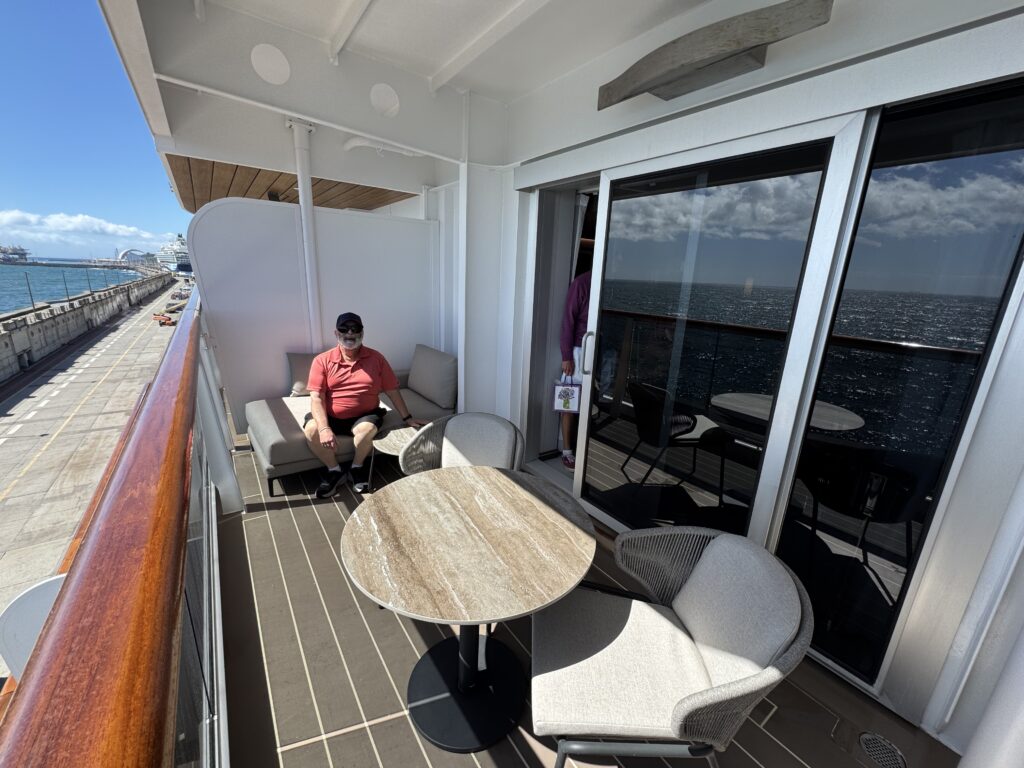 Man relaxing on a beige outdoor couch on a ship balcony, with round wooden table and glass doors to the interior, ocean and blue sky visible to the left.