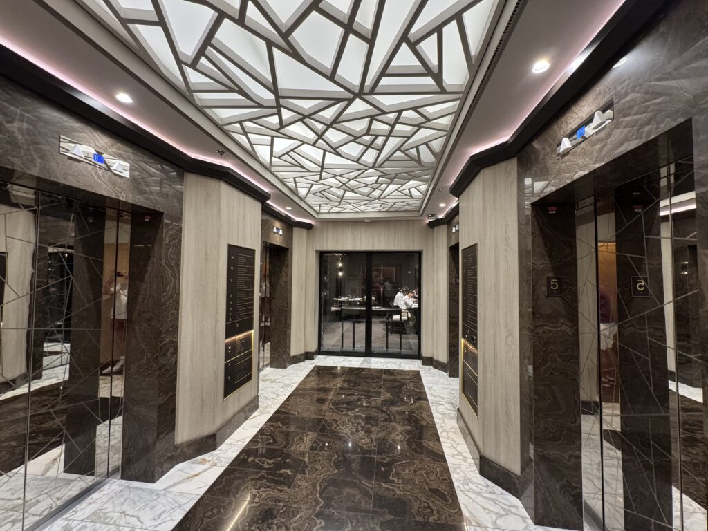 Interior of a modern elevator lobby with mirrored marble walls and glossy tiled floor, featuring a geometric skylight ceiling pattern and soft pink cove lighting along the walls at ceiling height