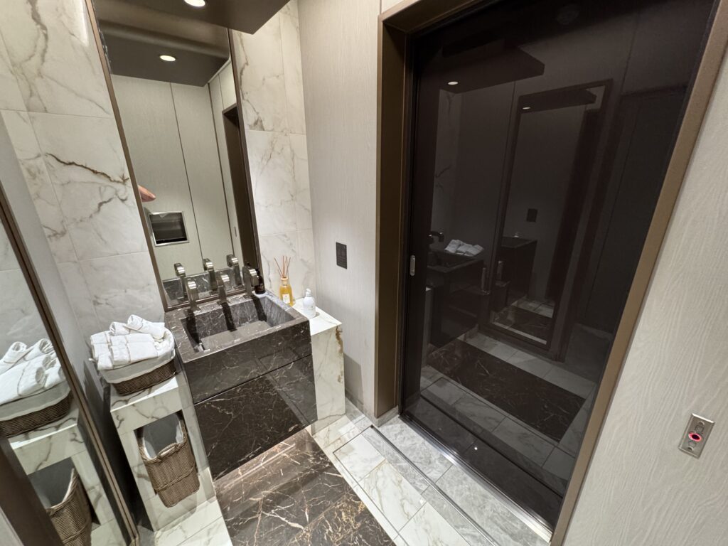 Hotel bathroom vanity with a black marble sink, chrome faucets, and marble walls; towels folded in baskets nearby.