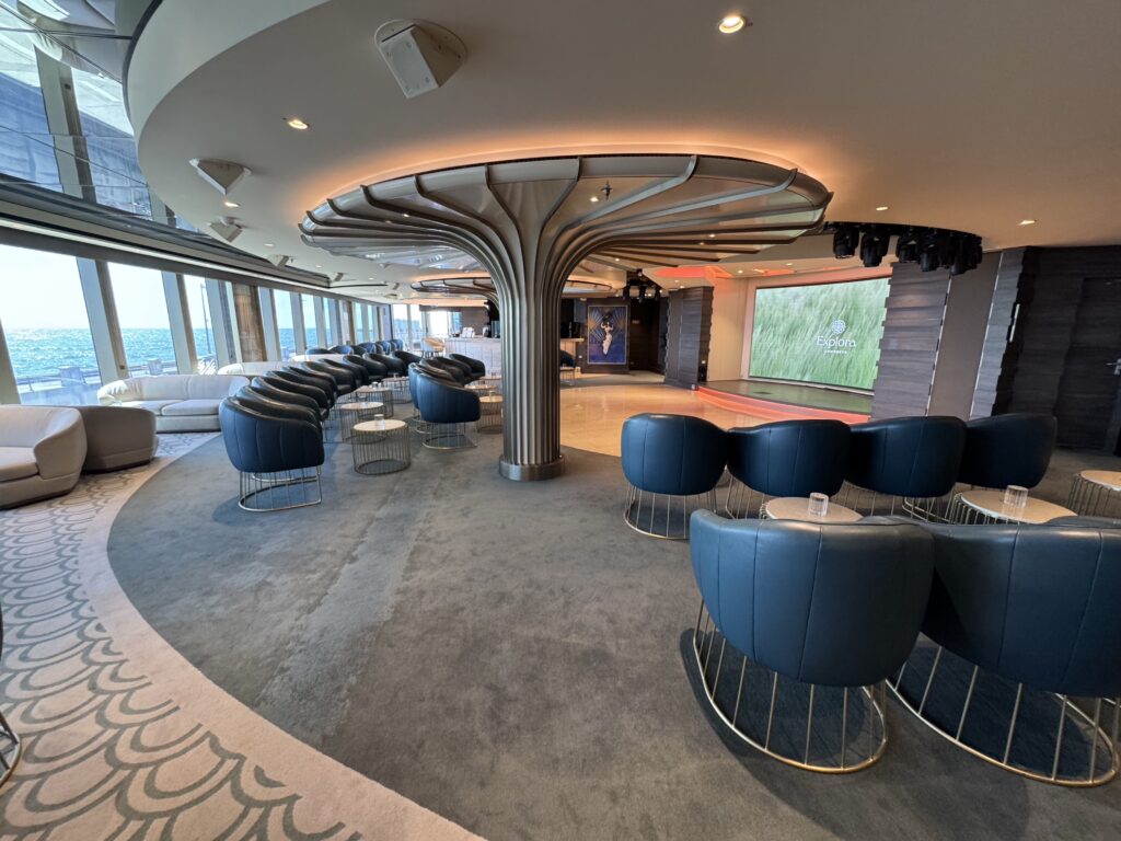 Cruise ship lounge with a sculptural central column, blue club chairs, circular tables, and a sea-view window wall.