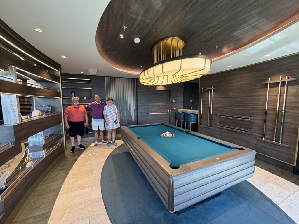 Three people stand and smile beside a blue-felt pool table in a modern billiards room with a large curved wood ceiling Light fixture above.