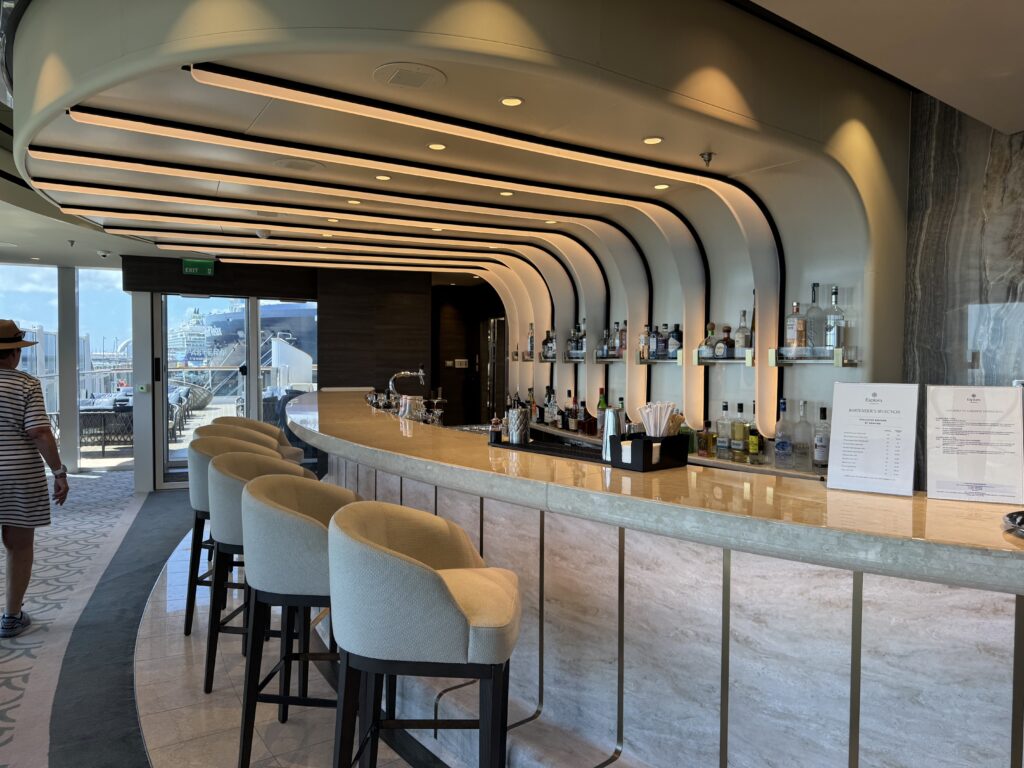 Modern bar interior with a curved white ceiling, horizontal light strips, and cream stools along a marble counter behind which liquor bottles are displayed.