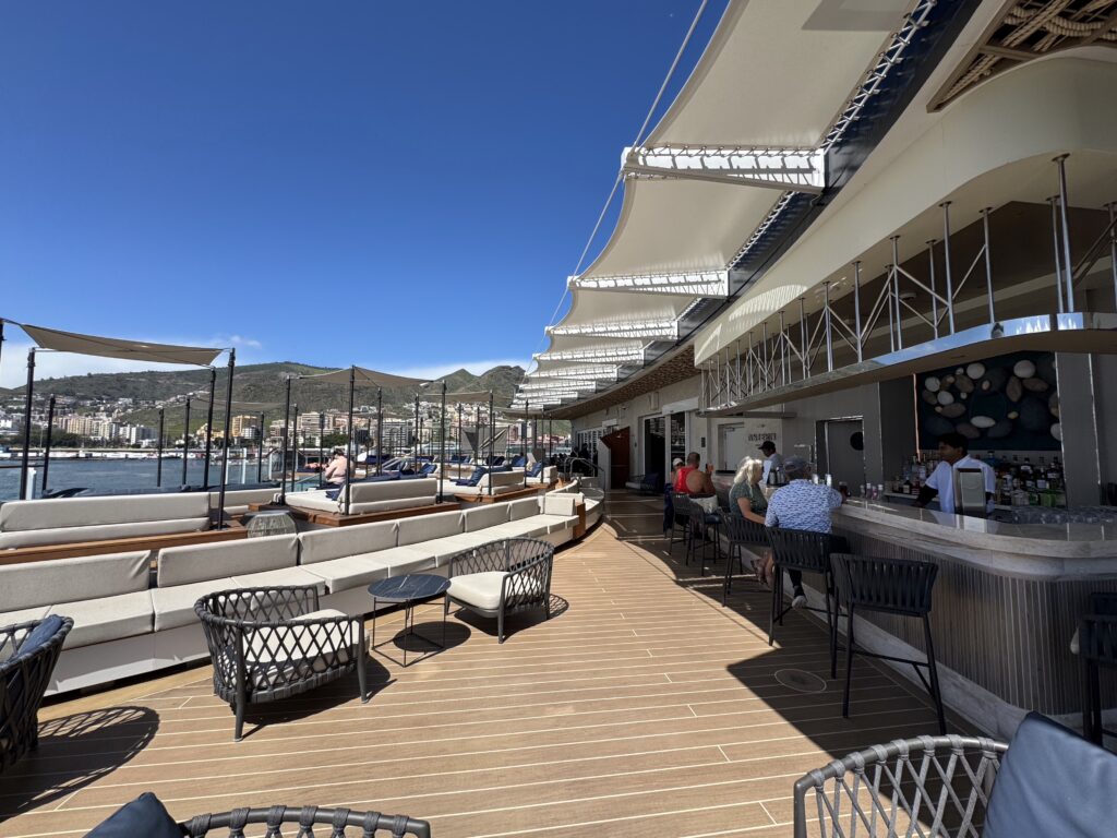Outdoor hotel bar and lounge area with wicker chairs, cushioned benches, and shade sails under a clear blue sky over a waterfront cityscape.