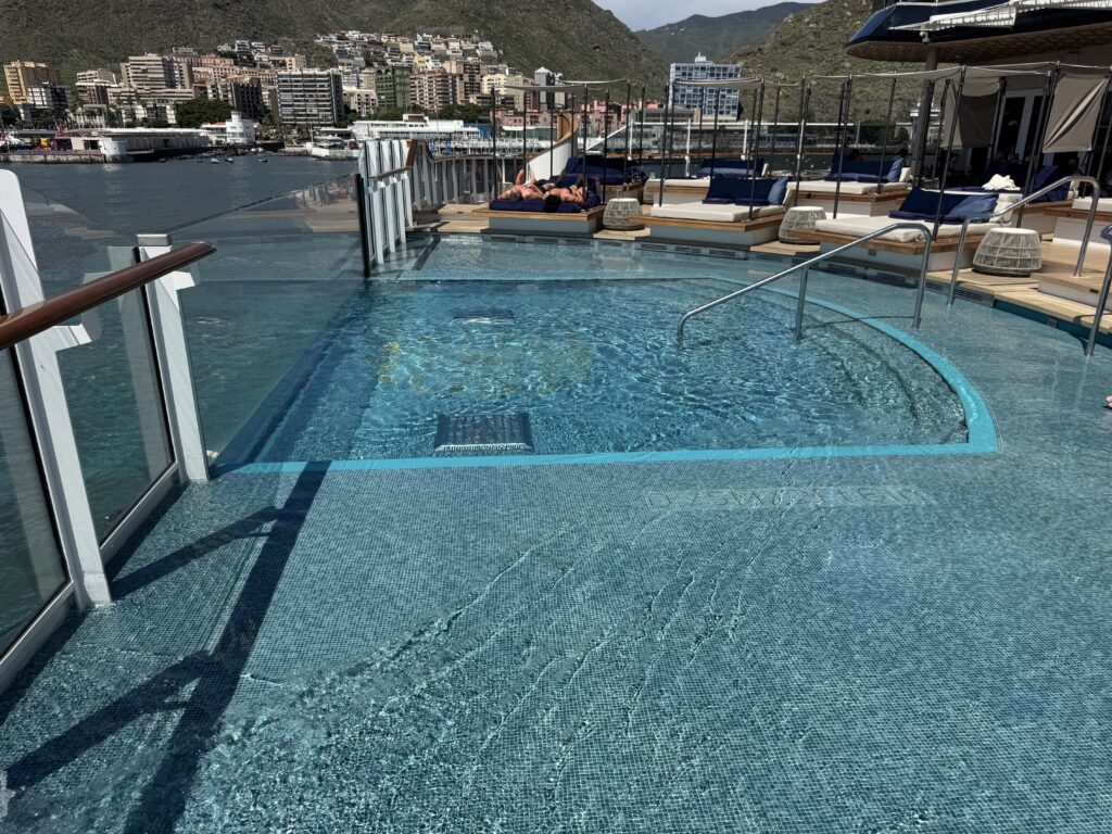 Sun loungers and couches on a rooftop pool deck, glass railings, with a city and hills in the background.